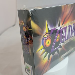 Pu&ograve; includere: Copia sigillata di The Legend of Zelda: Majora's Mask per Nintendo 64. La scatola presenta un design a maschera viola e gialla con il titolo del gioco. La scatola &egrave; sigillata in plastica e ha un logo N64 giallo.