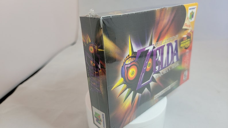Pu&ograve; includere: Copia sigillata di The Legend of Zelda: Majora's Mask per Nintendo 64. La scatola presenta un design a maschera viola e gialla con il titolo del gioco. La scatola &egrave; sigillata in plastica e ha un logo N64 giallo.