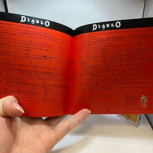 Peut inclure: Livre ouvert avec des pages rouges portant le titre "DIABLO" en blanc. Le livre semble &ecirc;tre un guide de jeu ou un manuel, avec des instructions et des d&eacute;tails sur le jeu.