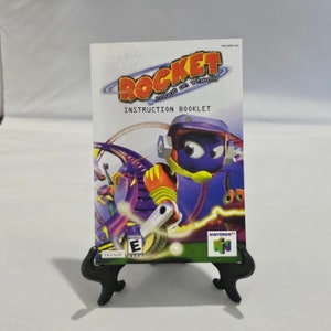 May include: A purple and yellow robot character on a purple and green track. The character is wearing a helmet and has a large, purple head. The image is from the instruction booklet for the Nintendo 64 game "Rocket: Robot on Wheels".