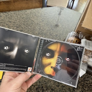 May include: Opened CD case featuring a close-up of a face with glowing eyes. The CD itself has a similar image. The case is held open, revealing the disc and the inside cover with a dark, shadowy image.