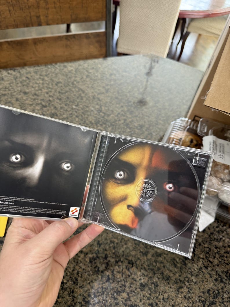 May include: Opened CD case featuring a close-up of a face with glowing eyes. The CD itself has a similar image. The case is held open, revealing the disc and the inside cover with a dark, shadowy image.