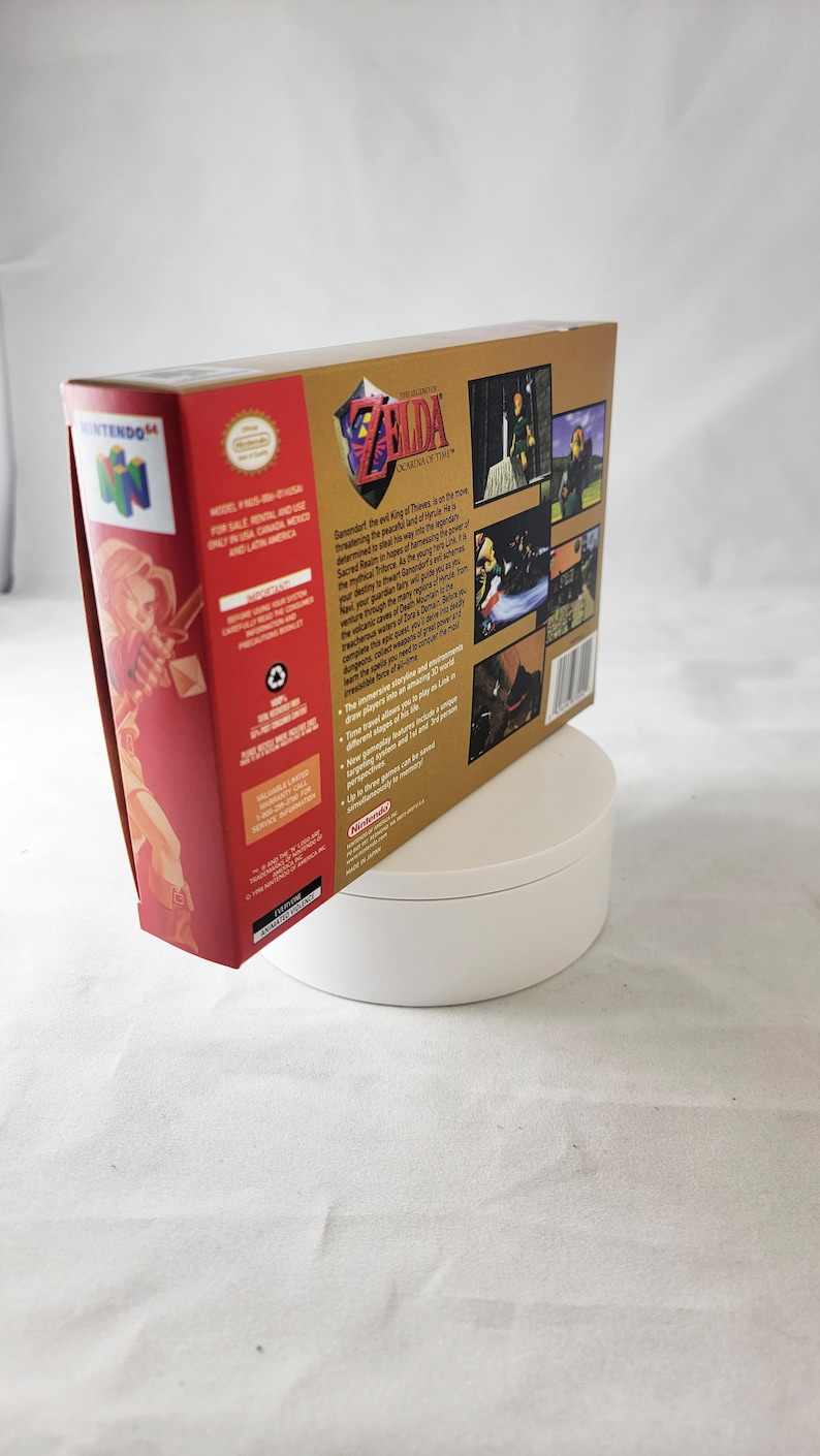 May include: A gold Nintendo 64 game cartridge case for The Legend of Zelda: Ocarina of Time. The front of the case features a red background with a drawing of Link holding a sword. The back of the case has a gold background with a description of the game and screenshots.