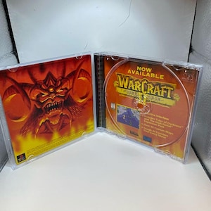 Peut inclure: Bo&icirc;tier de CD ouvert avec le jeu Warcraft: Dark Saga. Le CD pr&eacute;sente un design orange et marron avec le titre du jeu et du texte. L'illustration int&eacute;rieure du bo&icirc;tier repr&eacute;sente une figure d&eacute;moniaque rouge et orange. Le bo&icirc;tier est en plastique transparent.