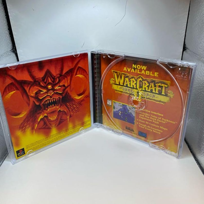 Peut inclure: Bo&icirc;tier de CD ouvert avec le jeu Warcraft: Dark Saga. Le CD pr&eacute;sente un design orange et marron avec le titre du jeu et du texte. L'illustration int&eacute;rieure du bo&icirc;tier repr&eacute;sente une figure d&eacute;moniaque rouge et orange. Le bo&icirc;tier est en plastique transparent.