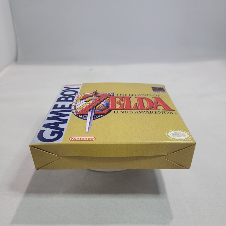 May include: A gold Game Boy cartridge case for The Legend of Zelda: Link's Awakening. The case has a blue Game Boy logo on the left side and a picture of Link with his sword and shield on the right side. The text "The Legend of Zelda: Link's Awakening" is printed in white on the case.