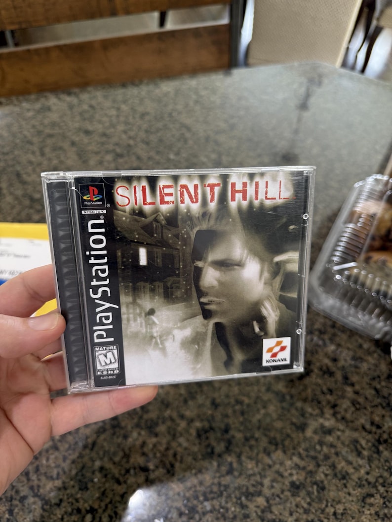 May include: A PlayStation game case for "Silent Hill." The cover art features a man's face, a house, and the game's title in red letters. The case is clear plastic, and the game is rated M for Mature.