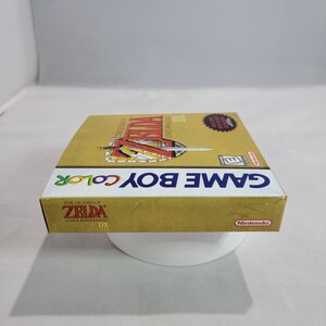 May include: A gold Game Boy Color game cartridge case for The Legend of Zelda: Link's Awakening DX. The case has a red and gold design with a sword and shield on the front. The text "GAME BOY COLOR" is printed in white on the front of the case.