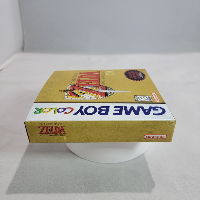 May include: A gold Game Boy Color game cartridge case for The Legend of Zelda: Link's Awakening DX. The case has a red and gold design with a sword and shield on the front. The text "GAME BOY COLOR" is printed in white on the front of the case.
