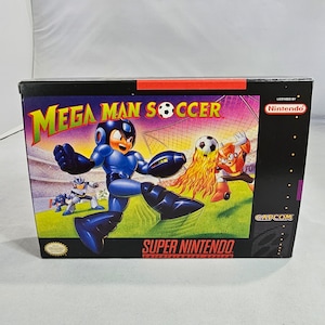 May include: A Super Nintendo Entertainment System game cartridge box for Mega Man Soccer. The box features a blue and black illustration of Mega Man kicking a football. The text "Mega Man Soccer" is in red and white letters. The box is black with a red and white Super Nintendo logo.