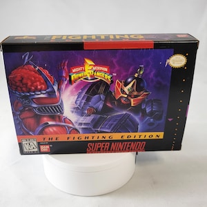 May include: A Super Nintendo game cartridge box for Mighty Morphin Power Rangers: The Fighting Edition. The box features a red and black Power Ranger fighting a black and silver robot. The box is black with red and yellow accents.