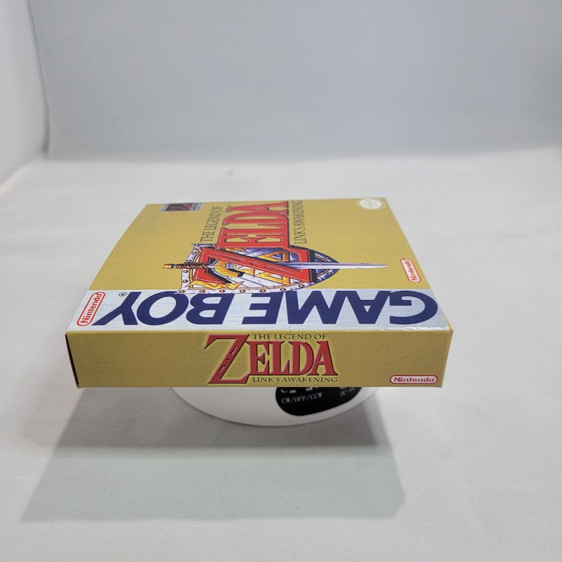 May include: A gold Game Boy cartridge case for The Legend of Zelda: Link's Awakening. The case has a picture of Link on the front with a sword and shield.