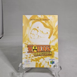 May include: A yellow instruction booklet for the Worms Armageddon video game for the Nintendo 64. The cover features a cartoon worm with a menacing expression and the game's title in bold letters. The booklet is made by Infogrames.