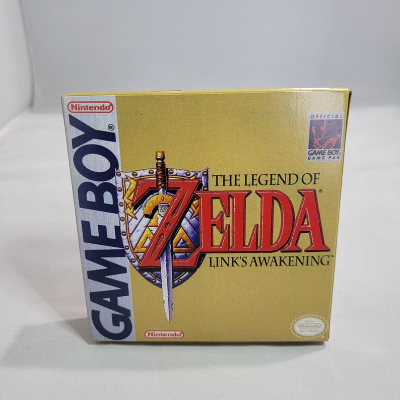 May include: A gold Game Boy cartridge case for The Legend of Zelda: Link's Awakening. The case features a red shield with a sword and the game title in white text.