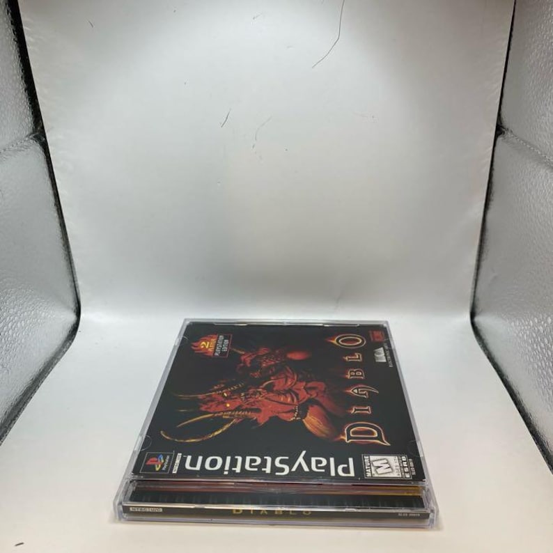 Peut inclure: Deux bo&icirc;tiers de jeu PlayStation empil&eacute;s. Le bo&icirc;tier sup&eacute;rieur pr&eacute;sente le jeu "Diablo" avec une illustration rouge et orange d'une figure d&eacute;moniaque. Le logo PlayStation et le titre du jeu sont visibles.