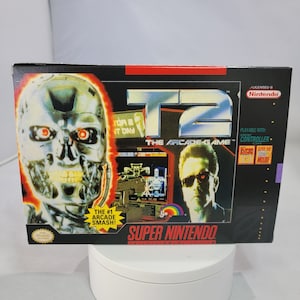 May include: A Super Nintendo game cartridge box for the game Terminator 2: Judgment Day - The Arcade Game. The box features a close-up image of the Terminator's face with red glowing eyes. The box also features a smaller image of Arnold Schwarzenegger as the Terminator from the movie. The text on the box reads "T2 The Arcade Game" and "Super Nintendo Entertainment System".