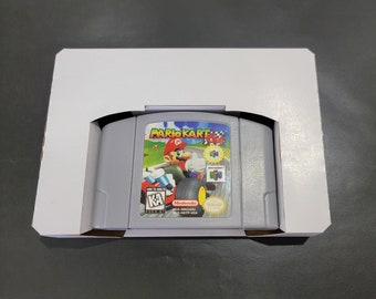 May include: A gray Nintendo 64 game cartridge with a white label. The label features a colorful image of Mario Kart 64, with Mario driving a kart on a race track. The text "Mario Kart 64" is printed in large, bold letters on the label. The game is rated "KA" for Kids to Adults.