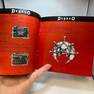Peut inclure: Livre ouvert intitul&eacute; "Diablo" avec des pages rouges. Des illustrations et du texte sur le jeu sont visibles. Une page affiche un dessin de cr&acirc;ne et d'os. Le livre est tenu ouvert par une main.