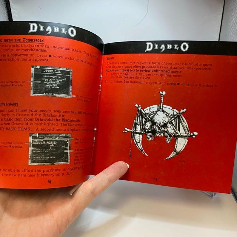Peut inclure: Livre ouvert intitul&eacute; "Diablo" avec des pages rouges. Des illustrations et du texte sur le jeu sont visibles. Une page affiche un dessin de cr&acirc;ne et d'os. Le livre est tenu ouvert par une main.