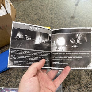 May include: Open book with black and white images and text. The title "PROLOGUE" is at the top. The images depict scenes from a video game. The text describes the story and characters. The book is held by a person.