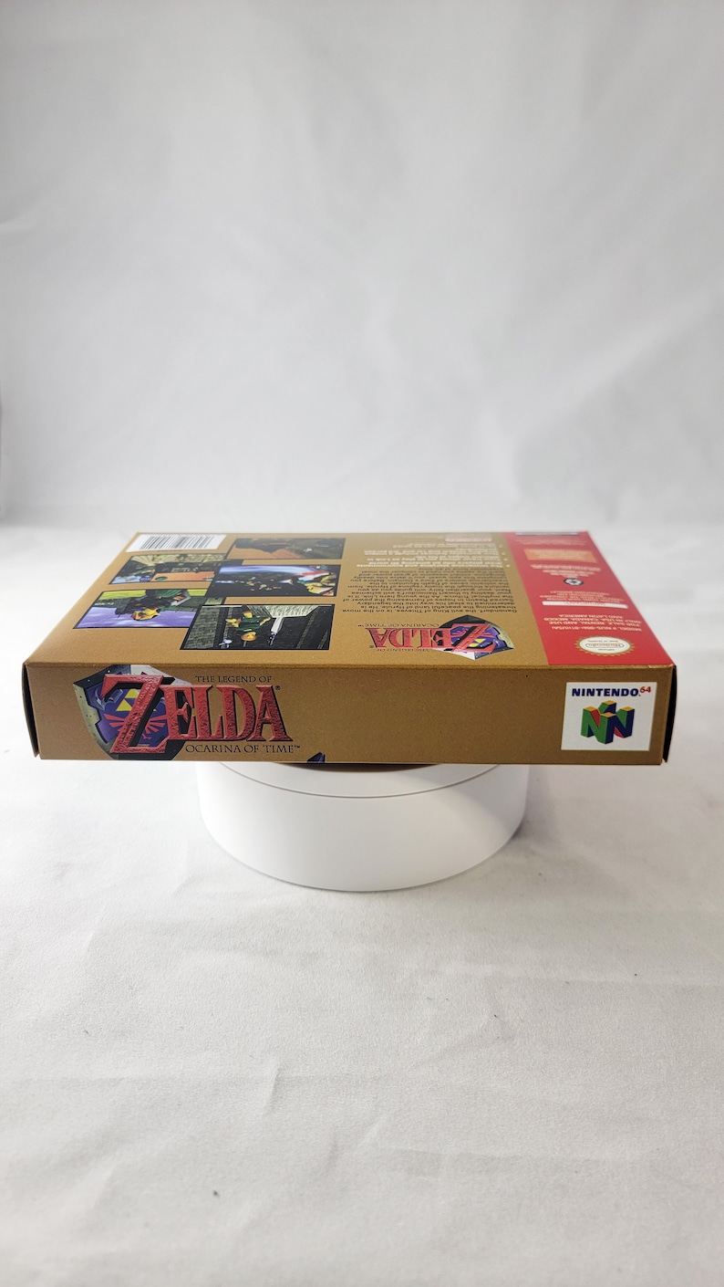May include: A gold Nintendo 64 game cartridge case for The Legend of Zelda: Ocarina of Time. The case has a picture of Link on the front and a picture of the game's logo on the back.