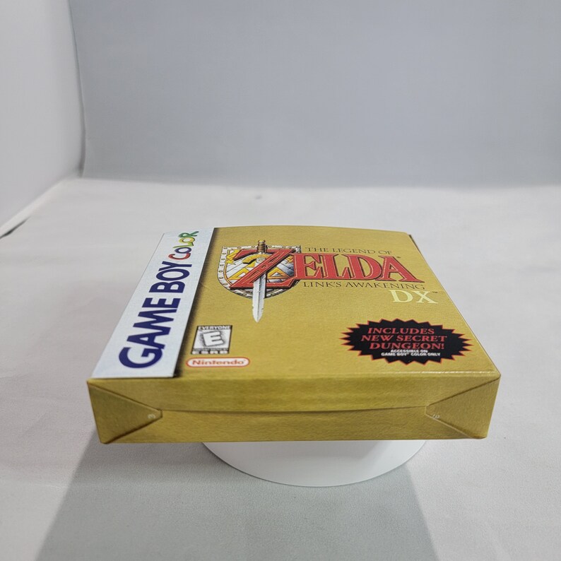 May include: A gold Game Boy Color game cartridge box for The Legend of Zelda: Link's Awakening DX. The box features a red and gold shield with a sword and the game title. The text "Includes New Secret Dungeon!" is on a red starburst.