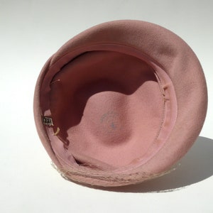 May include: A vintage pink cloche hat, viewed from the inside. The hat features a rounded crown and a narrow brim. A delicate, sheer veil is attached to the brim. The interior is lined with a matching pink fabric and a grosgrain ribbon.