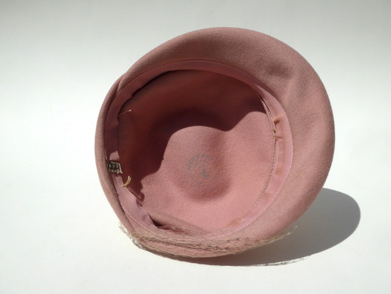 May include: A vintage pink cloche hat, viewed from the inside. The hat features a rounded crown and a narrow brim. A delicate, sheer veil is attached to the brim. The interior is lined with a matching pink fabric and a grosgrain ribbon.