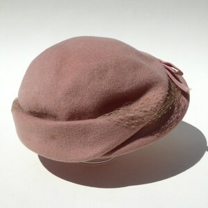 May include: A dusty rose cloche hat with a rounded crown and a turned-up brim. The hat features a small bow and a delicate netting detail. The hat is made of a soft, felt-like material.
