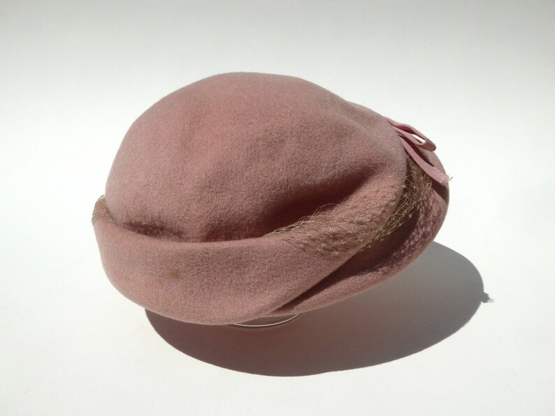 May include: A dusty rose cloche hat with a rounded crown and a turned-up brim. The hat features a small bow and a delicate netting detail. The hat is made of a soft, felt-like material.