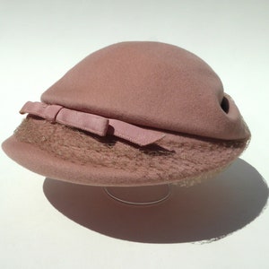 May include: A vintage pink cloche hat with a small bow and netting detail. The hat is made of a soft, felt-like material and is displayed on a clear stand. This hat is a classic accessory, perfect for adding a touch of vintage flair to any outfit.
