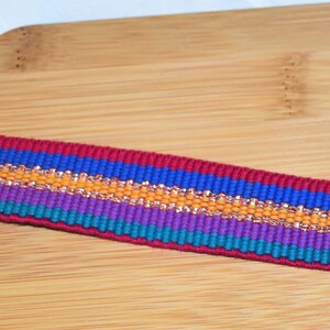 Puede incluir: Primer plano de una cinta tejida con rayas rojas, azules, moradas, verde azulado y doradas.