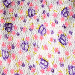 May include: White fabric with a repeating pattern of pink and purple flowers. The fabric has a textured surface.