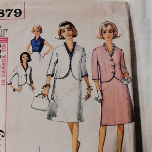 Op de afbeelding: Simplicity naaipatroon nummer 5879 voor een tienermaat 12T met een borstomvang van 81 cm. Het patroon is voor een ensemble van jas en rok. De afbeelding toont drie modellen die verschillende versies van de outfit dragen. Een model draagt een witte rok en jas met zwarte stippen, een ander draagt een roze rok en jas, en de derde draagt een blauwe top en een witte rok.