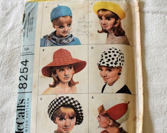 May include: A vintage sewing pattern for making hats. The pattern includes five different hat styles: a blue cloche, a red and white checkered beret, a yellow pillbox, a red and white pillbox, and a black and white polka dot pillbox. The pattern is titled "McCall's 8254 Printed Pattern" and is priced at 50 cents.