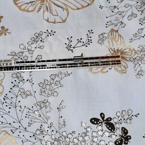 Pu&ograve; includere: Un tessuto bianco con un motivo a farfalle dorate e nere. Un righello con il testo "DRITZ" e "Sewing Gauge" &egrave; posizionato sul tessuto.