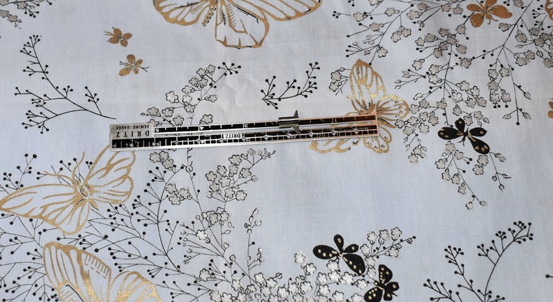 Pu&ograve; includere: Un tessuto bianco con un motivo a farfalle dorate e nere. Un righello con il testo "DRITZ" e "Sewing Gauge" &egrave; posizionato sul tessuto.