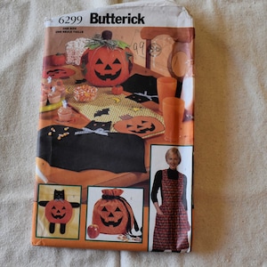 Könnte beinhalten: Ein Vintage-Butterick-Schnittmuster mit Halloween-Themen. Das Muster enthält Anleitungen zur Herstellung von Kürbisdekorationen, einem katzenförmigen Tischläufer und einer Schürze. Das Cover zeigt die fertigen Produkte in Orange, Schwarz und Gelb.