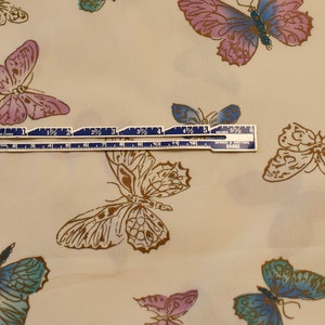 Pu&ograve; includere: Un tessuto beige con un motivo ripetitivo di farfalle di vari colori, tra cui blu, rosa e marrone. Un righello con segni in pollici &egrave; posizionato sopra il tessuto.