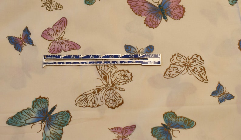Pu&ograve; includere: Un tessuto beige con un motivo ripetitivo di farfalle di vari colori, tra cui blu, rosa e marrone. Un righello con segni in pollici &egrave; posizionato sopra il tessuto.
