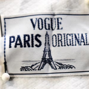May include: A white label with the words "VOGUE PARIS ORIGINAL" in dark blue. The Eiffel Tower is embroidered in the center. The label is pinned to a white fabric with small white ball pins.