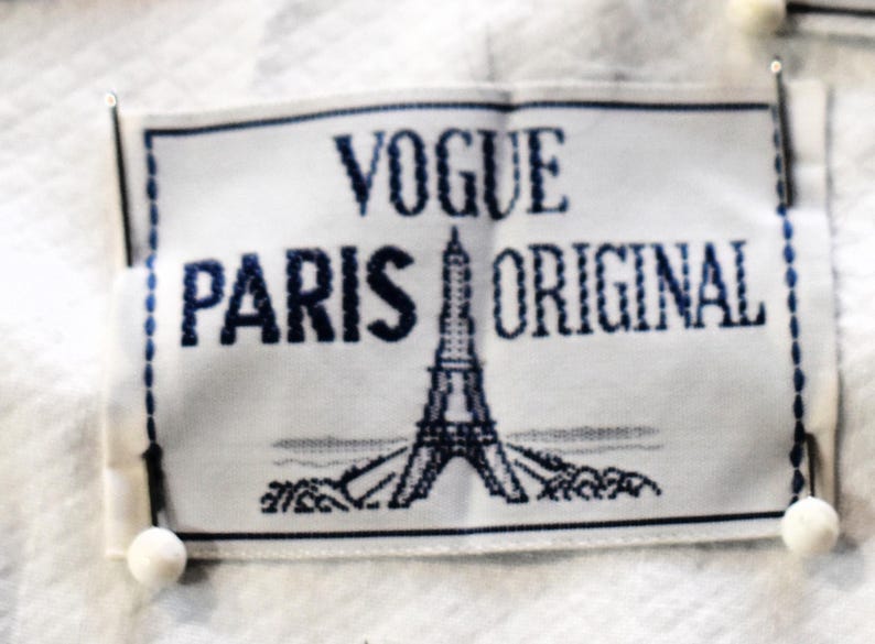 May include: A white label with the words "VOGUE PARIS ORIGINAL" in dark blue. The Eiffel Tower is embroidered in the center. The label is pinned to a white fabric with small white ball pins.