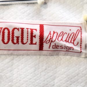 May include: A white fabric label with the words "VOGUE special design" in red lettering. The label is rectangular and pinned to a white textured fabric with black pins and white-headed pins.