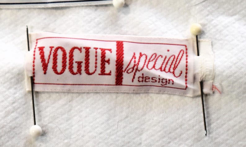 May include: A white fabric label with the words "VOGUE special design" in red lettering. The label is rectangular and pinned to a white textured fabric with black pins and white-headed pins.