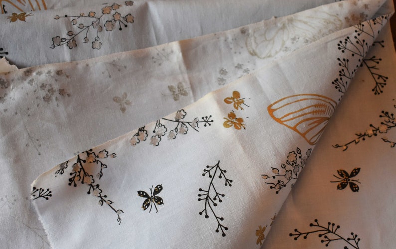 Pu&ograve; includere: Tessuto bianco con un motivo floreale e farfalle. Il tessuto presenta contorni neri di fiori e rami, farfalle dorate e piccoli fiori bianchi.