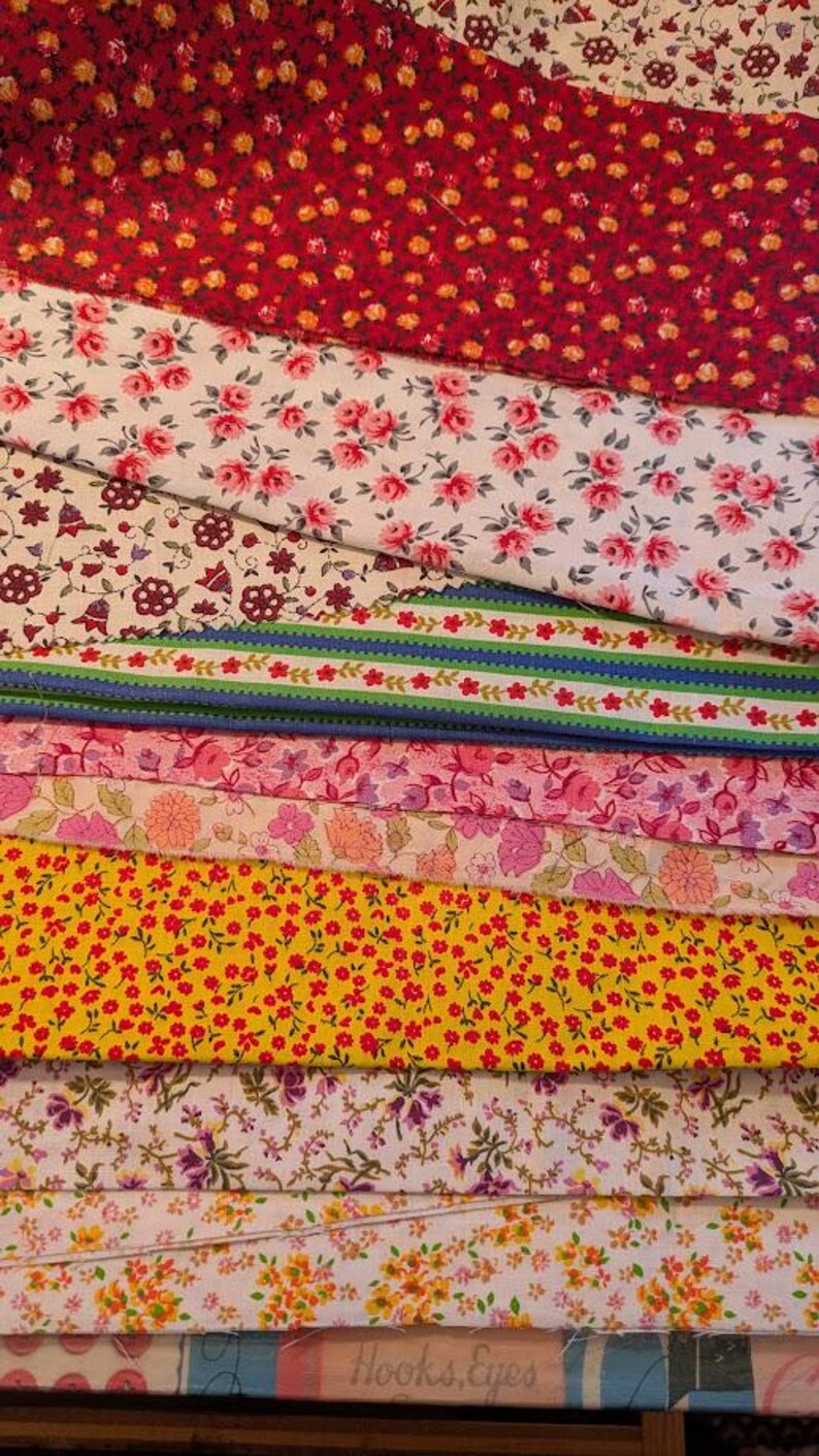 May include: A stack of various fabric swatches with floral patterns. The fabrics feature red, pink, yellow, and purple flowers on white, red, and yellow backgrounds. The text "Hooks, Eyes" is visible on the bottom fabric.