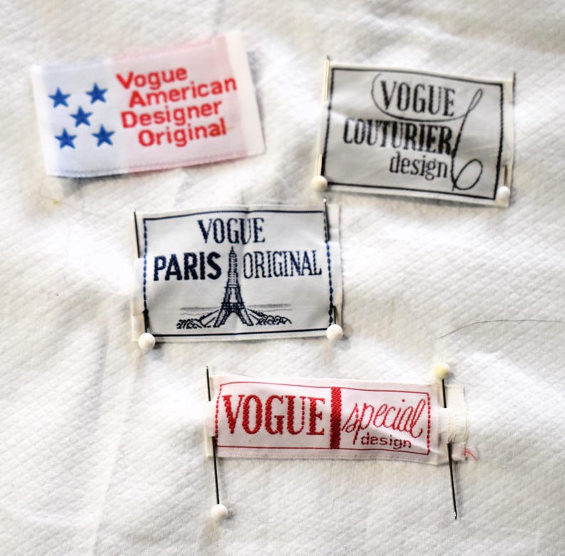 May include: Four fabric labels with the word "Vogue" printed on them. One label reads "Vogue American Designer Original" with blue stars. Another says "Vogue Couturier design". A third label says "Vogue Paris Original" with an Eiffel Tower illustration. The last label says "Vogue special design".