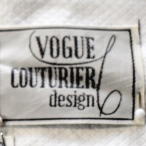 May include: A rectangular label with the text "VOGUE COUTURIER design" in black, set against a white background. The label has a black border and is slightly blurred. The label is attached to a white fabric.