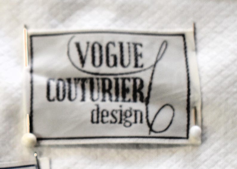 May include: A rectangular label with the text "VOGUE COUTURIER design" in black, set against a white background. The label has a black border and is slightly blurred. The label is attached to a white fabric.