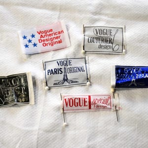 May include: A collection of vintage Vogue labels, each with unique text and designs. The labels feature phrases like "American Designer Original," "Paris Original," and "Special Design." The labels are pinned to a white textured surface.